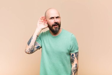 Genç kel ve sakallı bir adam gülümsüyor, tuhaf bir şekilde yan tarafa bakıyor, dedikoduları dinlemeye ya da bir sırrı duymaya çalışıyor.