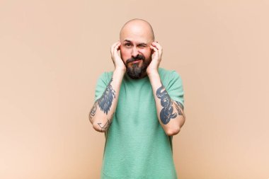 Genç kel ve sakallı adam sinirli, stresli ve sinirli görünüyor, iki kulağını da sağır edici bir ses, ses ya da yüksek sesle kapatıyor.