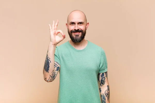 young bald and bearded man feeling happy, relaxed and satisfied, showing approval with okay gesture, smiling