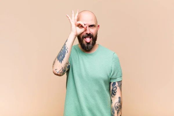 young bald and bearded man smiling happily with funny face, joking and looking through peephole, spying on secrets
