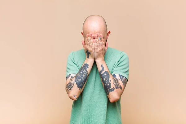 young bald and bearded man feeling sad, frustrated, nervous and depressed, covering face with both hands, crying