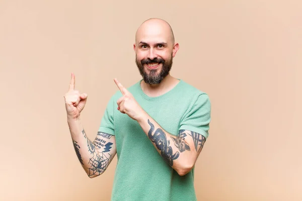 young bald and bearded man smiling happily and pointing to side and upwards with both hands showing object in copy space