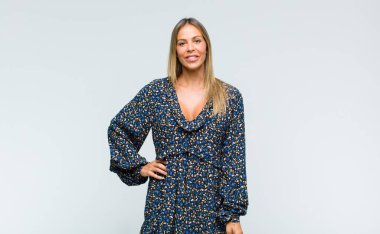 Genç ve güzel bir kadın mutlu bir şekilde gülümsüyor, eli kalçasında ve kendinden emin, pozitif, gururlu ve arkadaş canlısı.