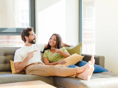 Genç çift neşeyle gülümsüyor, mutlu hissediyor ve yan ve yukarı doğru işaret ediyor, fotokopi uzayında nesneler gösteriyor. yeni ev kavramı