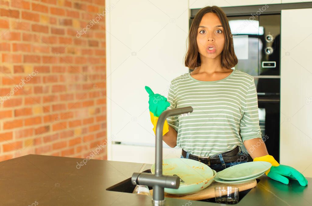 young pretty woman washing dishes looking astonished in disbelief, pointing at object on the side and saying wow, unbelievable