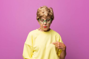 Yaşlı güzel kadın neşeyle gülümsüyor, yere bakıyor ve göğsüne işaret ediyor.