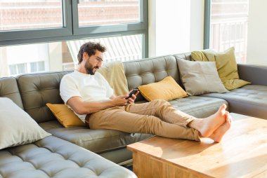 Genç sakallı havalı adam evdeki kanepede oturuyor.