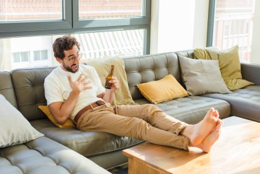 Genç sakallı havalı adam evdeki kanepede oturuyor.