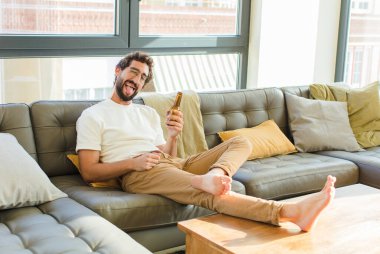 Genç sakallı havalı adam evdeki kanepede oturuyor.