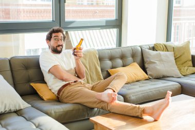 Genç sakallı havalı adam evdeki kanepede oturuyor.