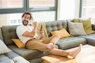 Genç sakallı havalı adam evdeki kanepede oturuyor.