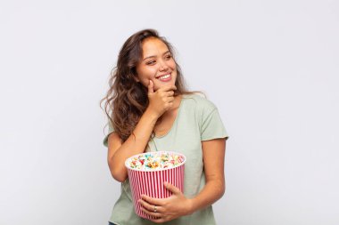 young woman with a pop conrs bucket smiling with a happy, confident expression with hand on chin, wondering and looking to the side