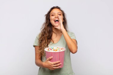 young woman with a pop conrs bucket feeling happy, excited and positive, giving a big shout out with hands next to mouth, calling out