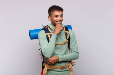 young hiker man smiling with a happy, confident expression with hand on chin, wondering and looking to the side