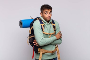 young hiker man shrugging, feeling confused and uncertain, doubting with arms crossed and puzzled look