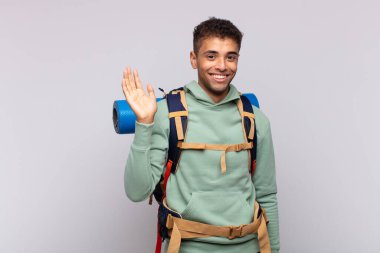 young hiker man smiling happily and cheerfully, waving hand, welcoming and greeting you, or saying goodbye