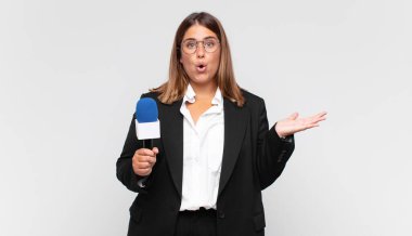 Genç kadın muhabir şaşırmış ve şoka uğramış görünüyor. Çenesi düşmüş ve eli açık bir objeyi tutuyor.