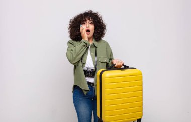 Genç Arap kadın şok olmuş ve korkmuş hissediyor, ağzı açık ve elleri yanaklarında seyahat konseptiyle dehşete düşmüş görünüyor.