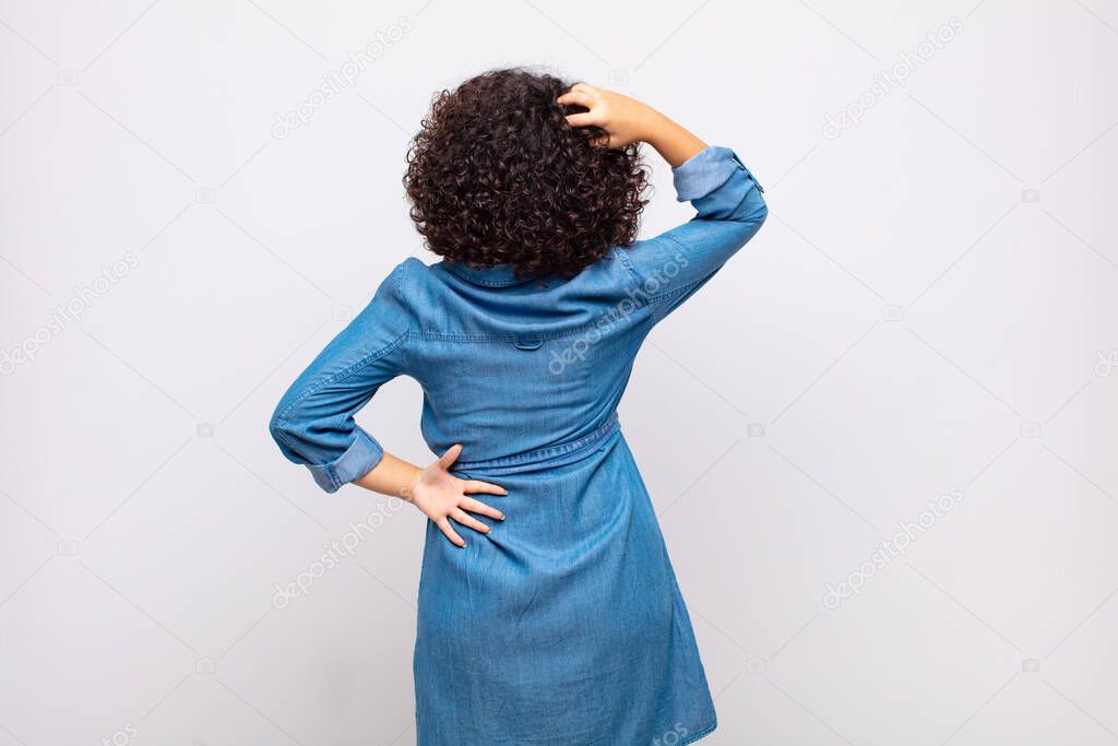 Woman feeling clueless and confused, thinking a solution, with hand on hip and other on head, rear view