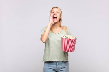 young woman with a pop corns bucket feeling happy, excited and positive, giving a big shout out with hands next to mouth, calling out
