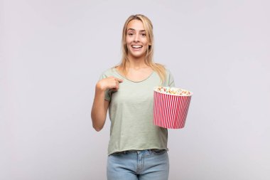 young woman with a pop corns bucket feeling happy, surprised and proud, pointing to self with an excited, amazed look