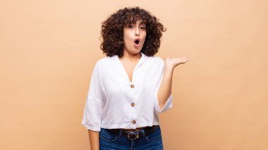 Kadın şaşırmış ve şok olmuş görünüyor, çenesi düşmüş ve eli açık bir objeyi tutuyor.