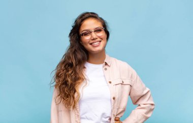 Kadın mutlu bir şekilde gülümsüyor, eli kalçasında ve kendinden emin, olumlu, gururlu ve arkadaş canlısı.