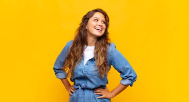 Mutlu, neşeli ve kendinden emin görünen, gururla gülümseyen ve iki elini de kalçasında taşıyan bir kadın.