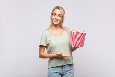 Elinde patlamış mısır kovası olan genç bir kadın neşeli bir şekilde gülümsüyor, mutlu hissediyor ve fotokopi uzayında avucunun içinde bir kavram gösteriyor.