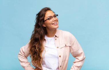 Mutlu, neşeli ve kendinden emin görünen, gururla gülümseyen ve iki elini de kalçasında taşıyan bir kadın.