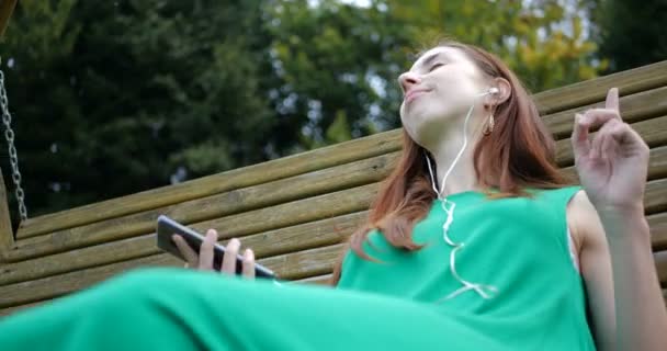 Baile emocional de la chica que escucha música con auriculares en el columpio . — Vídeos de Stock