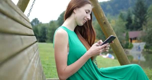 4k - chica sentada en el banco de oscilación y utilizar su teléfono en las montañas . — Vídeos de Stock