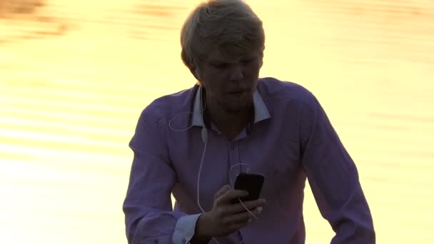 Homem loiro senta-se em uma margem do lago com caminho dourado do sol — Vídeo de Stock