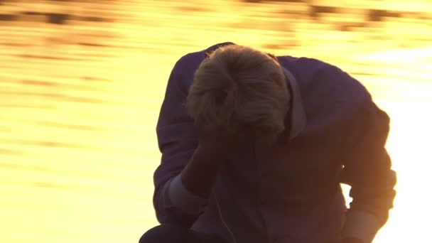 Νεαρός άνδρας ακούει μουσική σε μια τράπεζα στη λίμνη στο ηλιοβασίλεμα σε slo-mo — Αρχείο Βίντεο