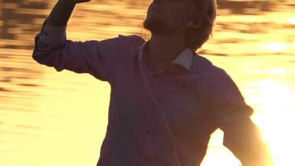 Jolly hombre escucha música en una orilla del lago al atardecer en slo-mo — Vídeo de stock
