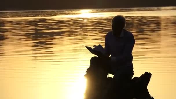 O jovem lê um livro em um banco de lago em um pôr do sol — Vídeo de Stock