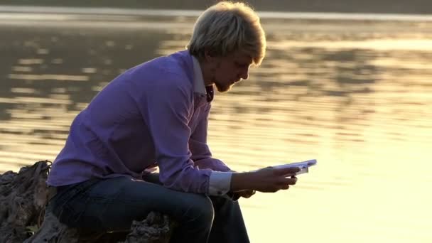 Estudiante se sienta en una orilla del lago y lee un manual — Vídeos de Stock