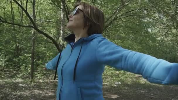 Medelålders kvinna med glasögon står i skogen och sprider sina armar. — Stockvideo