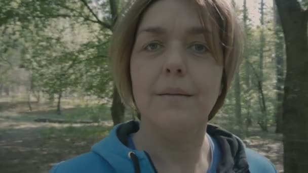Μεσήλικη γυναίκα στέκεται στο δάσος και tunrs με κάμερα και εμφάνιση. — Αρχείο Βίντεο