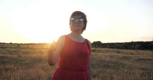 Preciosa mujer en gafas de sol y un vestido rojo bailando en un campo en slo-mo — Vídeo de stock