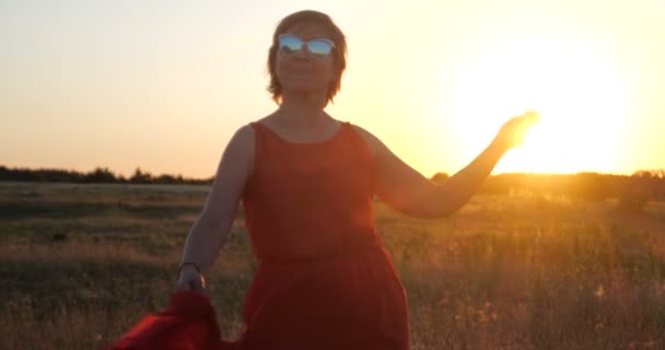 Sorrindo mulher elegante em um vestido vermelho dançando em um grande campo ao pôr do sol — Vídeo de Stock