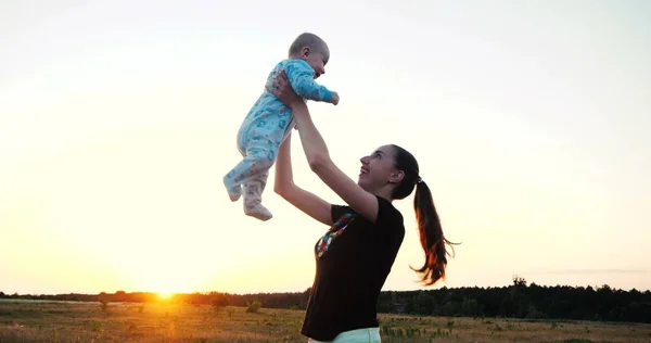 Felice madre con coda di cavallo che alleva il suo bambino in un grande campo al tramonto — Foto Stock