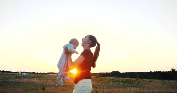 Емоційна молода жінка піднімає дитину в руках на полі на заході сонця — стокове фото