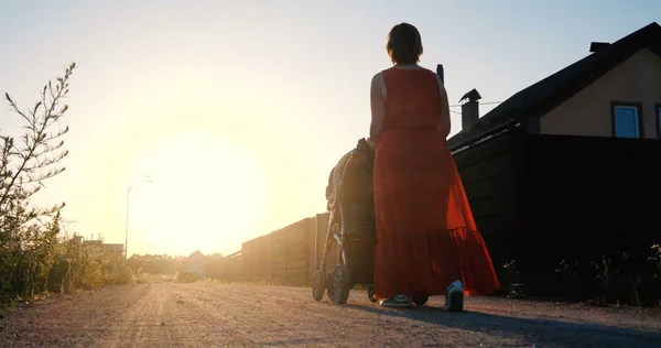 Slender woman in a red dress riding a baby in a pram at sunset — Stock Photo, Image