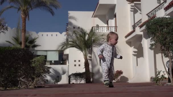 Un bambino cammina goffamente lungo una strada in una zona tropicale al rallentatore — Video Stock