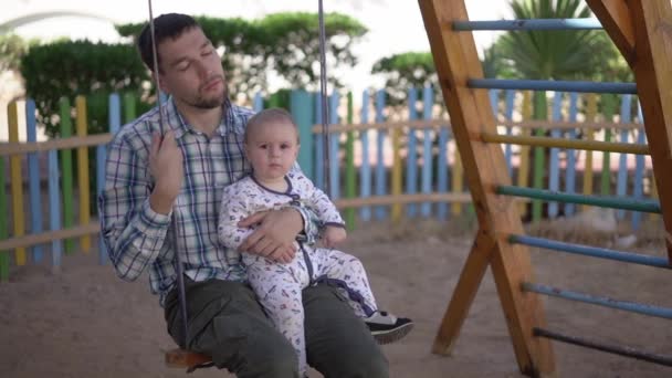 Trött pappa stänger sina Eys sitter på en gunga med en trött bebis i slow motion — Stockvideo