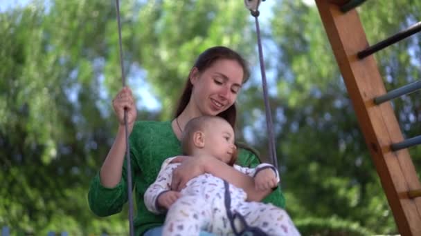 Mãe com uma criança de um ano de idade em seus braços montando um balanço em câmera lenta — Vídeo de Stock