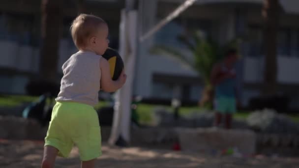 Милый мальчик берет волейбольный мяч на пляже и гуляет по пляжу . — стоковое видео