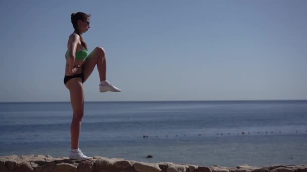 Slanke vrouw jogger maakt opwarmen van haar benen op het strand — Stockvideo