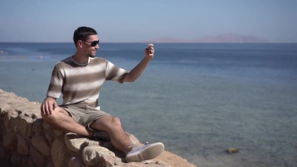 Jonge hipster op sunglases maakt foto van de Oceaan. — Stockvideo